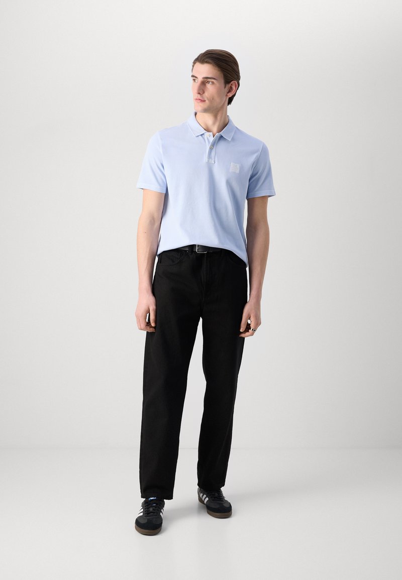 Helles blaues Poloshirt mit Kragen, versehen mit einem kleinen Logo, kombiniert mit schwarzen Hosen und dunklen Sneakers. Schlichte, moderne Passform.