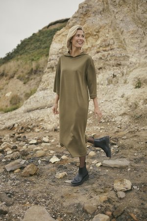Olivegrünes Kapuzenkleid mit drei Viertelärmeln, lockerer Silhouette und weicher Textur, kombiniert mit schwarzen Knöchelstiefeln, stehend auf steinigem Boden.