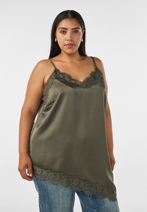 Kvinde med langt mørkt hår iført olivengrøn satin camisole med blondekant og blå jeans, stående mod en ensfarvet lys baggrund.