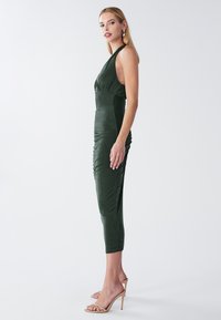 Obleka gozdno zelene barve s prileganim krojem, halterskim izrezom in naborki ob strani. Oblikovana je s srednje dolgo dolžino do meče.