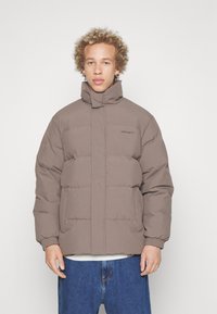 Brown puffer jacket with a high collar, elastic cuffs, and two front pockets; features a quilted design and subtle logo on the chest.