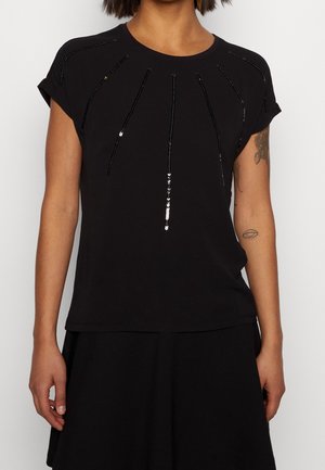 Kvinde iført sort kortærmet top med lodrette sorte pailletstriber, der stråler ud fra halsudskæringen, kombineret med sort nederdel.