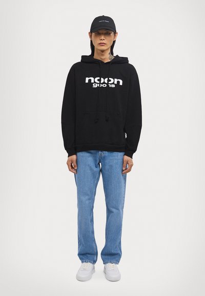 Schwarzer Kapuzenpullover mit weißem "noon goods" Logo, kombiniert mit hellblauen Jeans und weißen Sneakern. Eine schwarze Mütze rundet das Outfit ab.