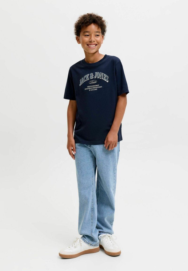 Garçon debout et souriant portant un t-shirt bleu marine Jack & Jones, un jean bleu clair et des baskets blanches sur un fond uni.