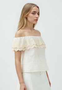 Bluzka typu off-the-shoulder w kremowym materiale z falbaniastą koronką, plisowanym wykończeniem u dołu oraz gładką strukturą.