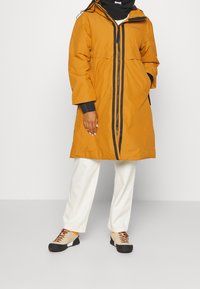 Orange vandtæt frakke med en sort lynlås, der har hætte, forlommer og en knælængde design. Kombineret med cremefarvede bukser og beige sko.
