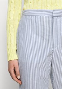 Žlutý svetr s pleteným vzorem u rukávu vedle světle modrých a bílých pruhovaných kalhot. Kalhoty mají hladkou texturu a plochý pas.