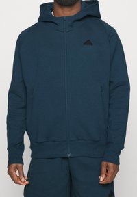 Man wearing zipped dark blue Adidas hoodie and matching shorts, standing with arms relaxed by sides, against a plain background.