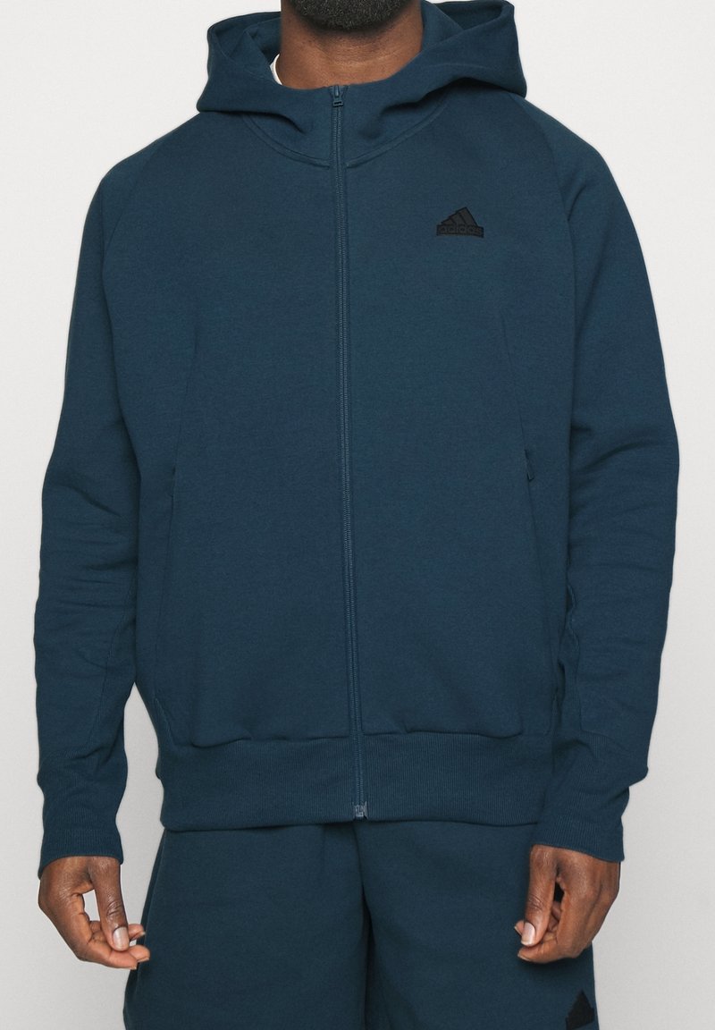 Man wearing zipped dark blue Adidas hoodie and matching shorts, standing with arms relaxed by sides, against a plain background.