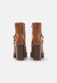 Brązowe skórzane botki za kostkę z teksturowanym tyłem, masywnym obcasem i ozdobnym wiązaniem z boku. Gładkie wykończenie z zaokrąglonym noskiem.