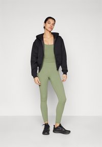 Crna zip-up hoodie, prilagođeni maslinasto zeleni top, usklađene tajice i crne atletske cipele. Glatka tkanina, ležeran dizajn i uski stil.