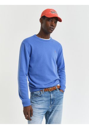 Pull en tricot bleu avec un col rond, un tissu texturé et des poignets côtelés ; associé à un jean bleu clair et une casquette de baseball rouge avec du texte.