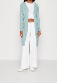 Jasnoniebieski kardigan z miękkiego materiału, z otwartym przodem, dwiema kieszeniami i długimi rękawami, w połączeniu z białymi spodniami w szerokim kroju i butami.