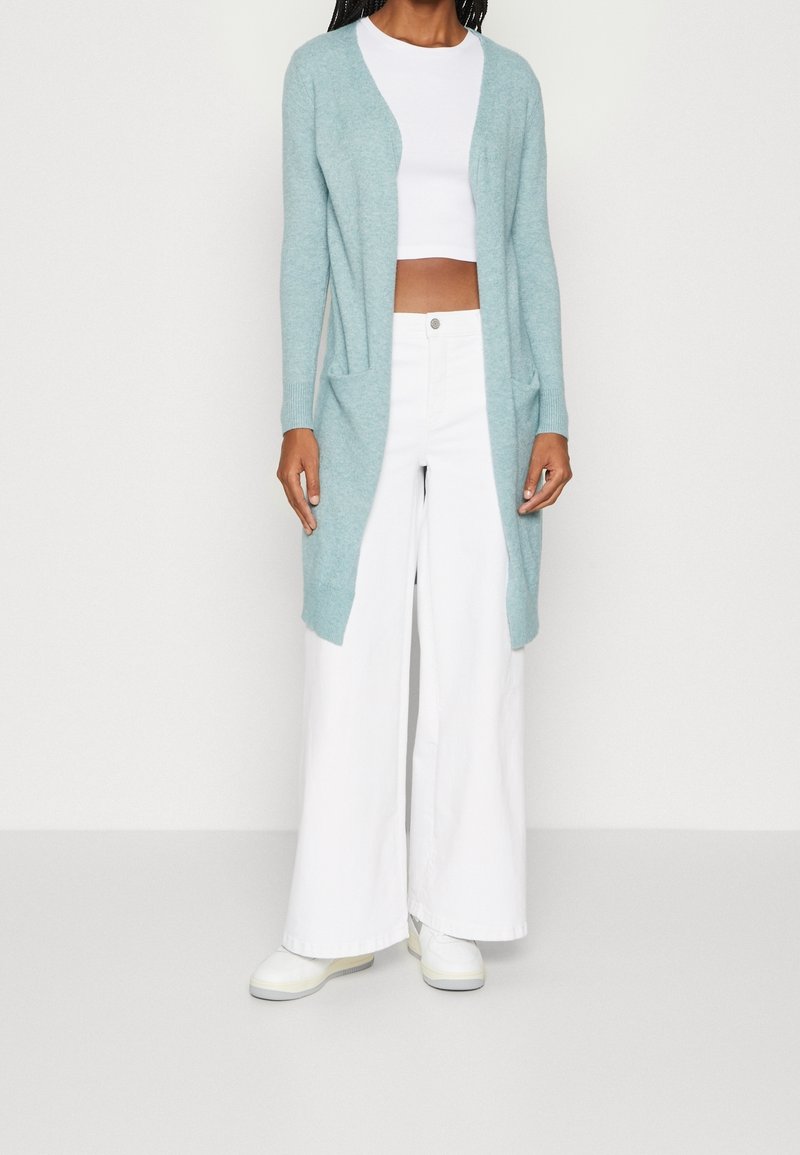 Jasnoniebieski kardigan z miękkiego materiału, z otwartym przodem, dwiema kieszeniami i długimi rękawami, w połączeniu z białymi spodniami w szerokim kroju i butami.