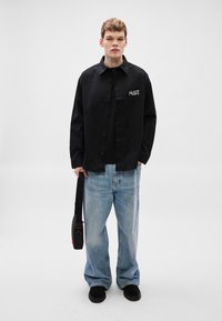 Μαύρο πουκάμισο με κουμπιά και λογότυπο στο μπροστινό μέρος, συνοδευόμενο από φαρδιά μπλε τζιν παντελόνια και μαύρα παπούτσια, κρατώντας μια μαύρη τσάντα χιαστί.