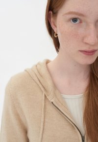 Ung person med långt rött hår som bär en beige zip-up hoodie och guldfärgade ringörhängen, med fräknar synliga i ansiktet och på halsen.