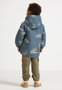 Bambino in pantaloni cargo verdi e stivali beige, che indossa una giacca blu con stampe di dinosauri, visto di spalle su uno sfondo bianco uniforme.
