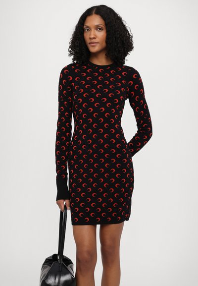 Vestido de punto con base negra y patrones en forma de media luna rojos, con mangas largas y una silueta ajustada, acompañado de un bolso negro.