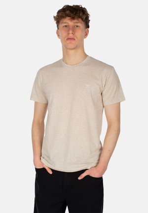 Beige T-Shirt mit kurzen Ärmeln aus einem weichen, strukturierten Stoff, mit Rundhalsausschnitt und einem kleinen weißen Logo auf der linken Brust. Getragen mit dunklen Hosen.