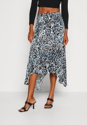 Frau trägt einen schwarz-weißen, abstrakten, asymmetrischen Midirock, ein schwarzes, bauchfreies Langarmtop und schwarze Sandalen mit Absatz.