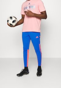 Růžové tričko značky Juventus s reflexním logem, v kombinaci s modrými sportovními kalhotami s růžovými pruhy. Černé fotbalové kopačky.