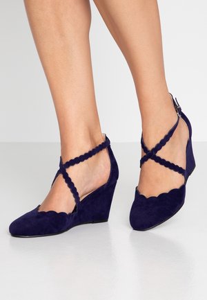 Escarpins compensés en daim bleu marine avec bords festonnés et brides croisées à la cheville. Dotés d'un bout pointu et d'une surface texturée.