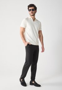 Weißes kurzärmliges Poloshirt mit gerippter Textur, kombiniert mit schwarzen Slim-Fit-Hosen und schwarzen Wildlederschuhen, Sonnenbrille rundet den Look ab.