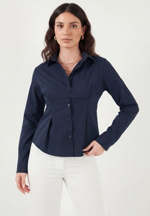 Femme aux longs cheveux ondulés portant une chemise ajustée bleu marine à boutons et un pantalon blanc, accessoirisée avec des boucles d’oreilles créoles dorées et des colliers superposés.