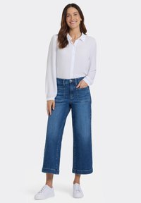 Vit långärmad skjorta med knappstängning, kombinerad med vida blå jeans i denim. Modellen bär vita sneakers och har händerna i fickorna.