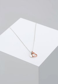 Zilveren ketting met twee in elkaar grijpende hartpendanten; de ene is zilver en de andere is roségoud. Vlekkeloze afwerking, modern design.