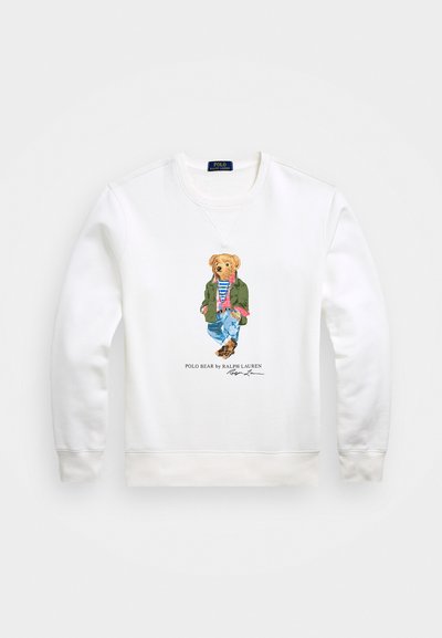 Hvit hettegenser med en brodert bjørn som har på seg en grønn jakke og blå jeans, med en stripete skjorte og brune sko.