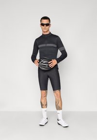 Cycling outfit includes a black long-sleeve top with gray stripes, black shorts, and white shoes. Model holds a black helmet.