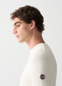 Suéter de punto color crema hecho de material suave; presenta un cuello redondo y un parche de logo redondo en la manga izquierda.