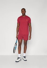 Camiseta e calções atléticos vermelhos, feitos de tecido leve. Apresentam um pequeno logótipo na camisola. Usados com ténis de ténis e uma raquete.