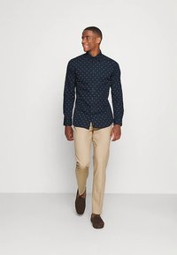 Marineblaue Hemdbluse mit weißen Punkten, langen Ärmeln und einem normalen Kragen, kombiniert mit beigen Hosen und braunen Schuhen.