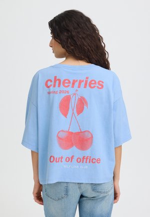 Femme aux longs cheveux bouclés porte un t-shirt bleu ample avec des cerises rouges et le texte « cherries spring 2026 Out of office SELF LOVE CLUB » dans le dos.