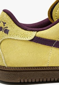 Sneaker giallo in suede con accenti e cuciture viola. Suola in gomma testurizzata con un motivo rigato. Presenta una punta rotonda e un collare imbottito.
