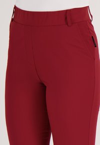 Close-up van een persoon die een strak, hooggetailleerd rood broek draagt met voor- en zijzakken en een wit shirt.