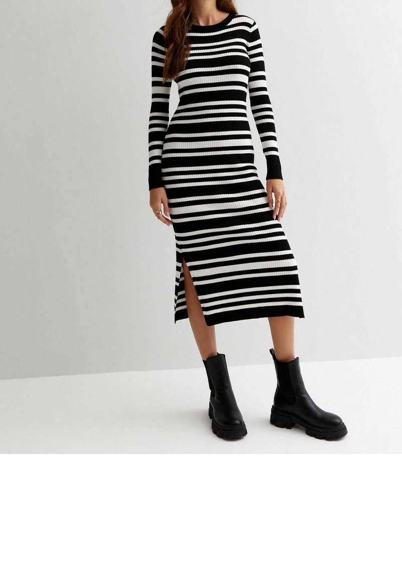 Femme portant une robe midi à manches longues rayée horizontalement en noir et blanc avec une fente latérale et des bottines noires épaisses.