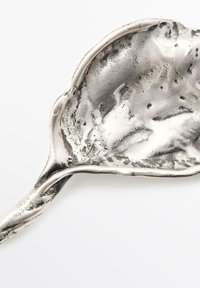 Silbernes, metallisches blattförmiges Geschirr mit einer strukturierten Oberfläche und glatten Kanten, ausgestattet mit einem verlängerten, gebogenen Griff.