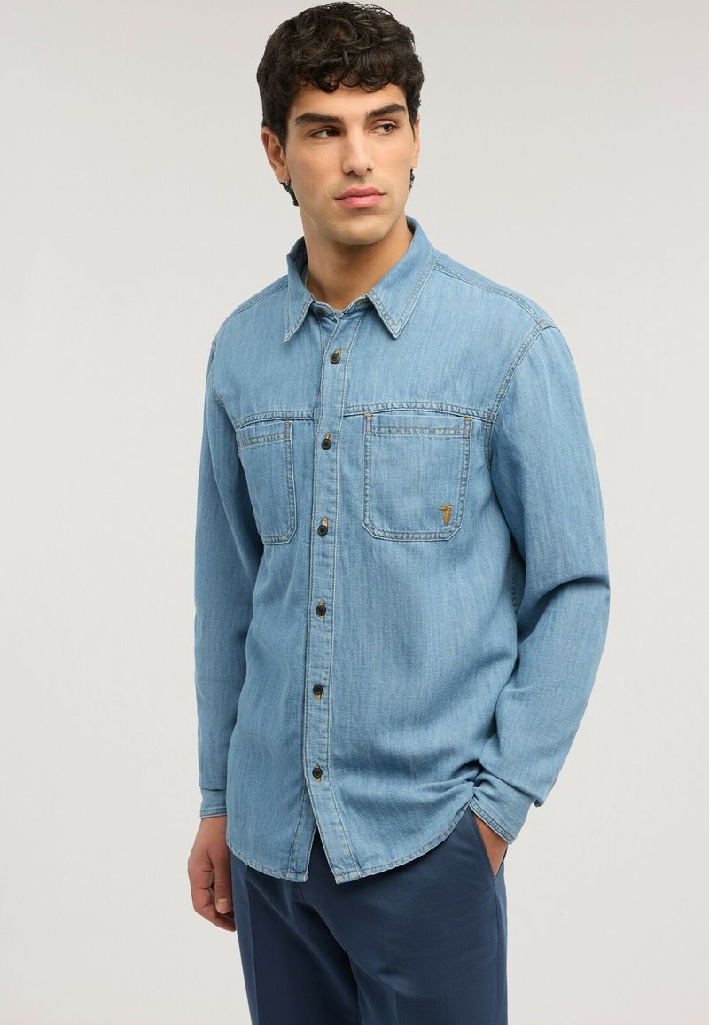 Joven con cabello rizado oscuro, lleva una camisa de mezclilla azul claro abotonada y pantalones oscuros, posando con una mano en el bolsillo.