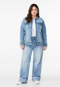 Šviesiai mėlynas džinsinis švarkas su mygtukų uždarymu priekyje, dviem krūtinės kišenėmis ir derančiais laisvalaikio šviesiai mėlynais džinsais. Audinys atrodo švelnus ir dėvėtas.