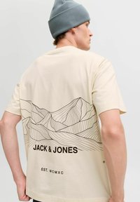 Beež T-särk, mille seljal on musta joonejoonistuse kujutis mägedest. Allosas on "JACK & JONES" logo ja "EST. MCMXC" tekst.