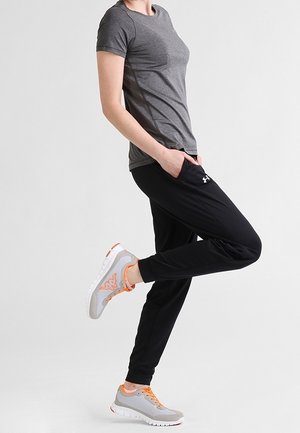 Persona che indossa una maglietta grigia, pantaloni jogger neri e scarpe da ginnastica grigie con lacci arancioni, in piedi su una gamba con le mani nelle tasche.