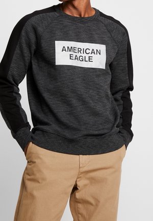 Osoba ubrana w ciemnoszarą i czarną bluzę z napisem "AMERICAN EAGLE" na przodzie oraz beżowe spodnie, z rękami w kieszeniach.