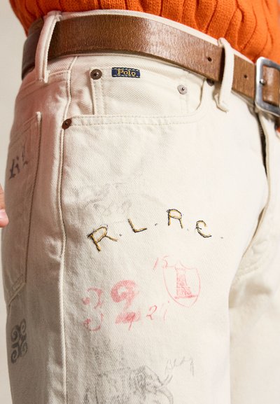 Hvite bukser med "Polo"-merke og påsydde bokstaver "R.L.R.C.", brukt med brunt skinnbelte og oransje strikkegenser.