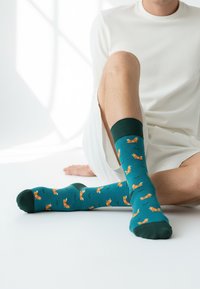 Tirkizne nogavice z oranžnimi vzorci lisice, temno zelenim robom, gladko teksturo in podaljšanim dizajnom do kolen.