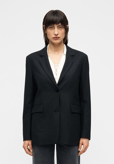 Blazer noir sur mesure à un seul bouton, avec des revers nets et deux poches avant, confectionné dans un tissu lisse avec une silhouette structurée.