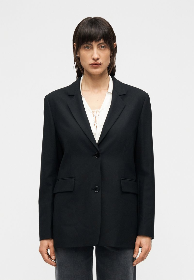 Μαύρο ραμμένο σακάκι με ένα κουμπί, κοφτερά πέτα  γιακά και δύο μπροστινές τσέπες, κατασκευασμένο από λείο ύφασμα με δομημένη σιλουέτα.