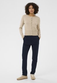 Beez värviga kudum kaartigan ümmarguse kaeluse ja nööpide ees, koos tumesiniste pükstega ja heleda beeži tossudega. Sile tekstuur, keha külge liibuv mudel.